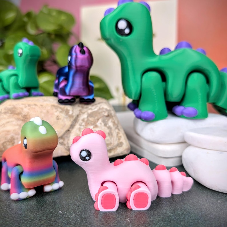 Op de afbeelding: Een verzameling kleurrijke, beweegbare dinosaurusfiguren. De dinosaurussen zijn in verschillende kleuren, waaronder groen, roze en regenboogtinten. Elke figuur heeft grote, expressieve ogen en een speels ontwerp.