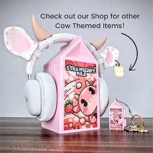 May include: Pink cow-themed headphones with a pink milk carton stand. The milk carton has a cartoon cow and the words "Strawberry Milk" on it. There is a smaller pink milk carton keyring with a cow on it.