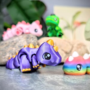 May include: A collection of colorful, 3D-printed dragon figurines. The foreground features a purple dragon with gold accents. Other dragons are pink, green, and rainbow-colored. Each dragon has large, expressive eyes, and is posed on a natural stone surface.