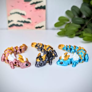 K&ouml;nnte beinhalten: Drei bewegliche Drachenfiguren in Rosa, Schwarz und Gold sowie Blau mit goldenen Akzenten. Jeder Drache hat ein detailliertes Design mit Fl&uuml;geln und einem segmentierten K&ouml;rper. Die Drachen werden auf einer wei&szlig;en Oberfl&auml;che pr&auml;sentiert.