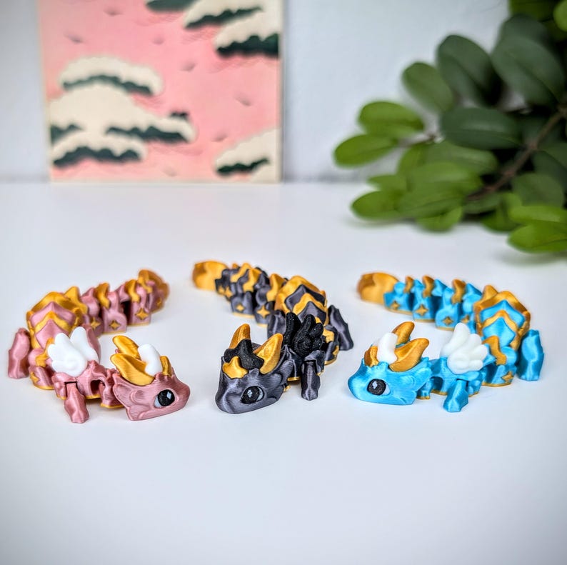 K&ouml;nnte beinhalten: Drei bewegliche Drachenfiguren in Rosa, Schwarz und Gold sowie Blau mit goldenen Akzenten. Jeder Drache hat ein detailliertes Design mit Fl&uuml;geln und einem segmentierten K&ouml;rper. Die Drachen werden auf einer wei&szlig;en Oberfl&auml;che pr&auml;sentiert.