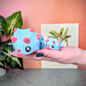 May include: Two blue and pink dinosaur figurines. The larger dinosaur is standing on its four legs and has a pink nose and eyes. The smaller dinosaur is sitting on its hind legs and has a pink nose and eyes.