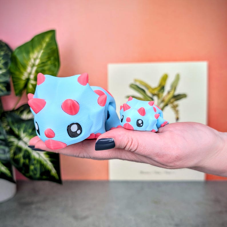 May include: Two blue and pink dinosaur figurines. The larger dinosaur is standing on its four legs and has a pink nose and eyes. The smaller dinosaur is sitting on its hind legs and has a pink nose and eyes.