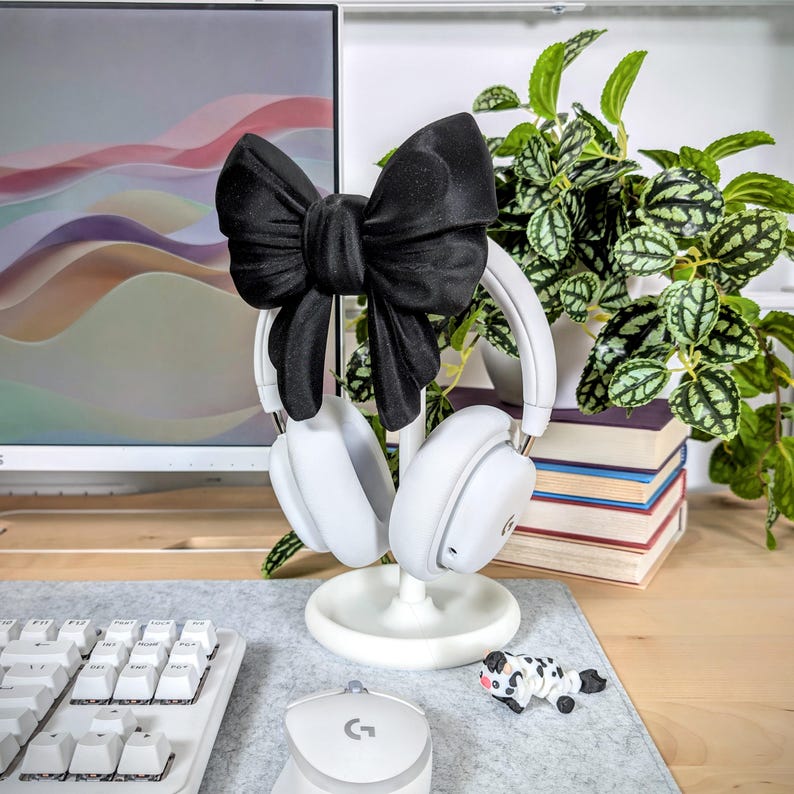 May include: White headphones with a black bow accent rest on a white stand. A white keyboard and mouse are in the foreground. A small cow figurine and a plant are also visible. The background includes a computer monitor and a wooden desk.