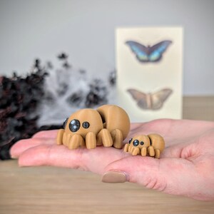 May include: Two tan-colored, 3D-printed spider figurines, one large and one small, are displayed. The spiders have large, cartoonish eyes. The larger spider is approximately 2 inches long. A blurred background includes a card with butterfly illustrations.