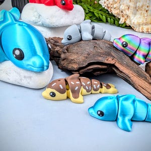 May include: A collection of colorful, articulated dolphin toys in various shades of blue, red, gray, brown, yellow, and rainbow. The toys are arranged on white stones and a piece of driftwood, with a seashell in the background.