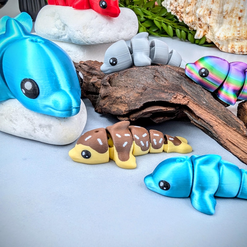 May include: A collection of colorful, articulated dolphin toys in various shades of blue, red, gray, brown, yellow, and rainbow. The toys are arranged on white stones and a piece of driftwood, with a seashell in the background.
