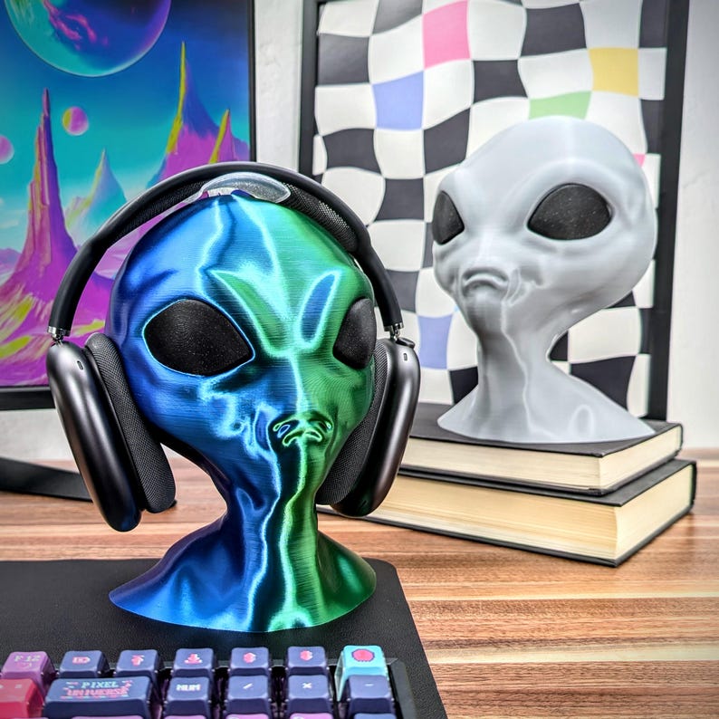 May include: A green and blue alien head headphone stand with a pair of black headphones on it. The stand is on a wooden table next to a gray alien head stand.