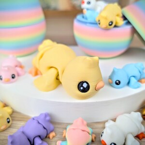 May include: A collection of colorful, miniature animal figurines, including yellow, blue, pink, and purple creatures. The figurines have large, dark eyes and small beaks. Some are lying down, while others are in a rainbow-colored container.