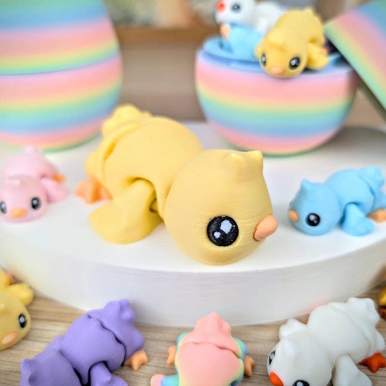 May include: A collection of colorful, miniature animal figurines, including yellow, blue, pink, and purple creatures. The figurines have large, dark eyes and small beaks. Some are lying down, while others are in a rainbow-colored container.
