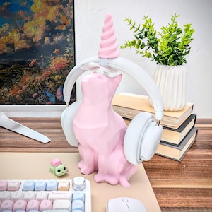 May include: A pink cat-shaped headphone stand with a unicorn horn on top. The stand is holding a pair of white over-ear headphones. The stand is on a wooden surface with a keyboard and a mouse.