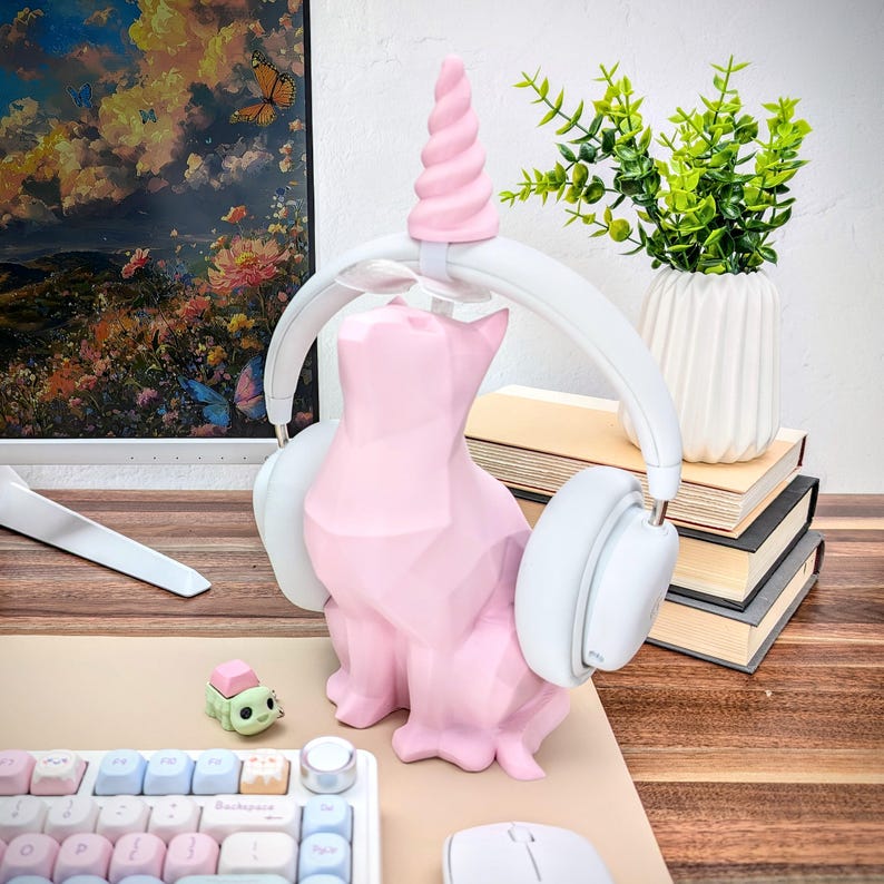 May include: A pink cat-shaped headphone stand with a unicorn horn on top. The stand is holding a pair of white over-ear headphones. The stand is on a wooden surface with a keyboard and a mouse.
