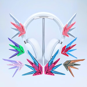 May include: White headphones with colorful, wing-shaped attachments in various colors, including pink, blue, green, and orange. The attachments have a geometric design and are displayed with the headphones on a stand.