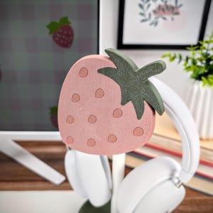 May include: A pink and green strawberry-shaped headphone stand. The stand is made of plastic and has a textured surface. The stand is designed to hold headphones.