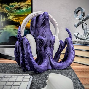 May include: Purple octopus-shaped headphone stand with a white pair of headphones resting on its head. The stand is made of 3D printed plastic.