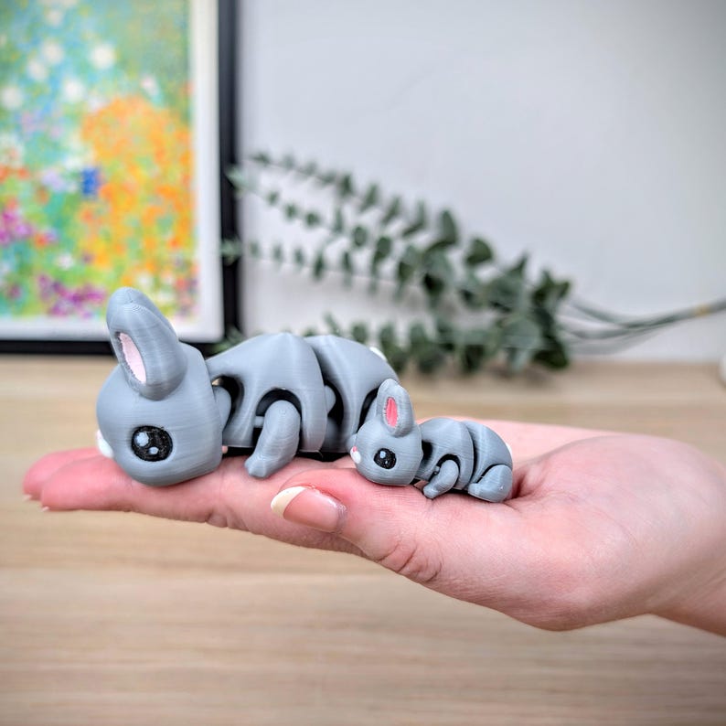 May include: Three grey articulated bunny figurines with black eyes and pink inner ears are displayed. The bunnies are of varying sizes, with the largest in the front and the smallest in the back. The bunnies are held in a hand.