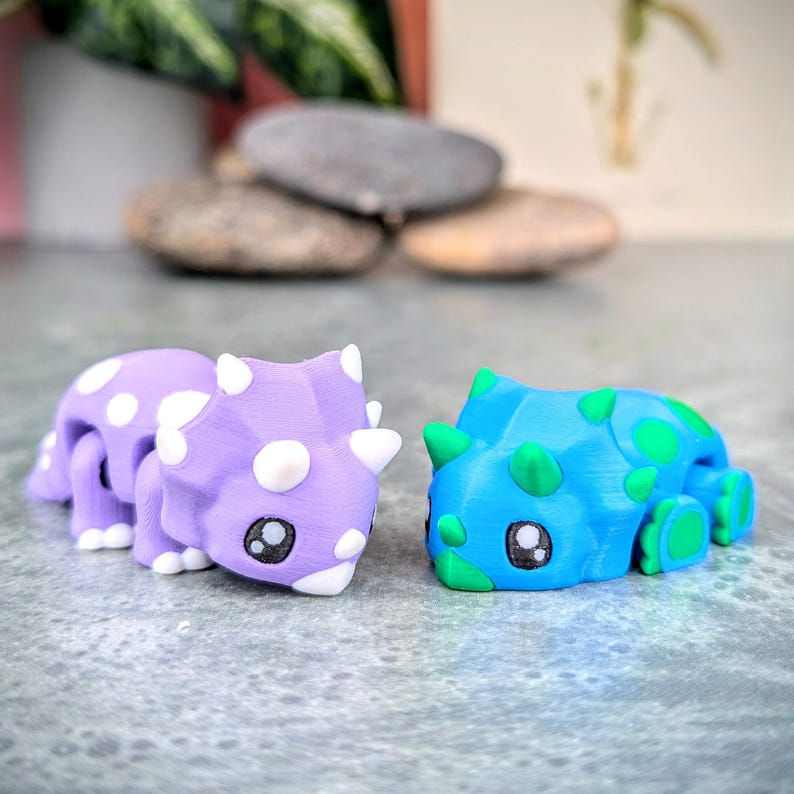 May include: Two small, cartoon-like dinosaur figurines. One is purple with white spots and the other is blue with green spots.