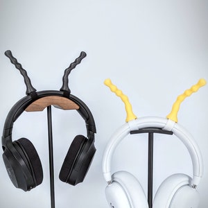 May include: Two pairs of headphones on a stand. The headphones on the left are black with a wooden stand. The headphones on the right are white with a black stand. Both headphones have a yellow or black rubber hook on the top.