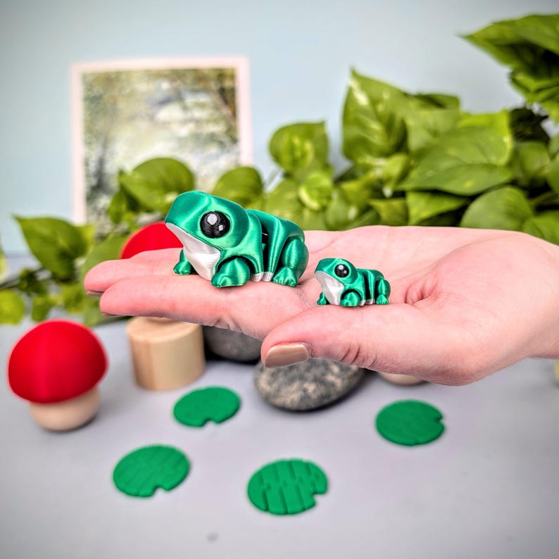 May include: Two green frog figurines with large black eyes and white accents. A larger frog and a smaller frog are displayed. The image also includes a red mushroom, wooden cylinders, green lily pads, and small stones.
