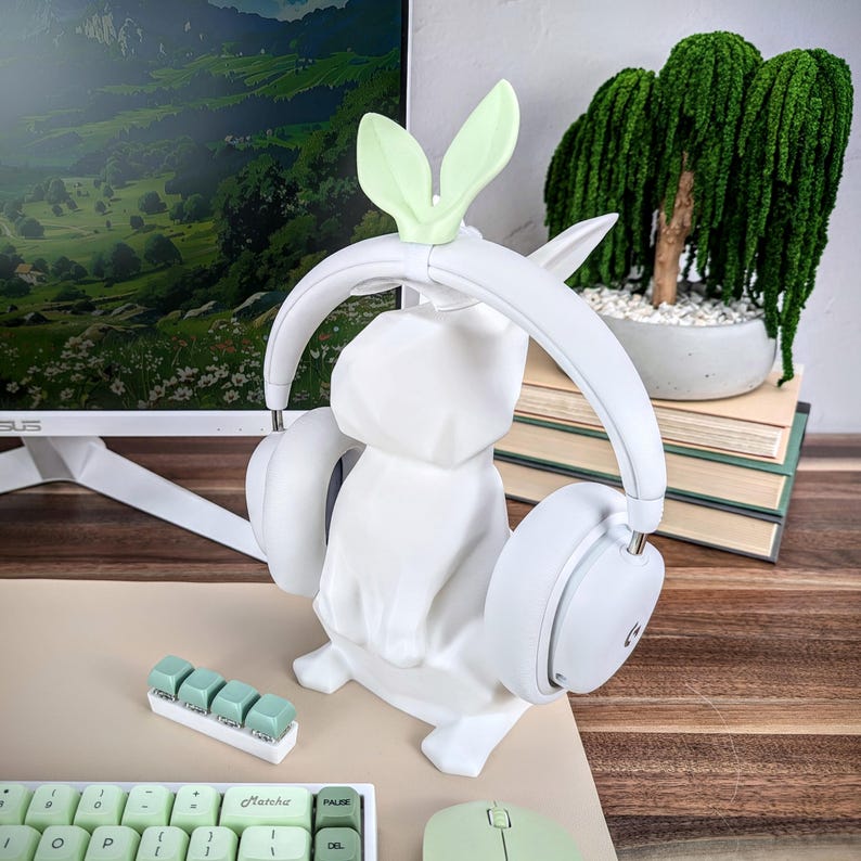 May include: White bunny-shaped headphone stand with green bunny ears. The stand is holding a pair of white headphones. The stand is on a wooden desk with a keyboard and mouse.
