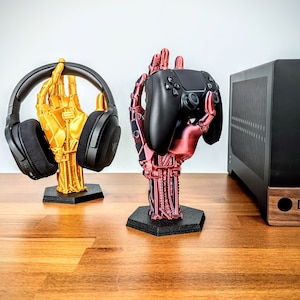May include: Two robotic hand-shaped stands, one gold and one red, hold a black headset and a black game controller. The stands have a detailed mechanical design and sit on black hexagonal bases. A black computer case is in the background.