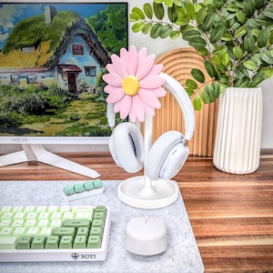 May include: White headphone stand with a pink daisy flower design. The stand is holding a pair of white headphones. The stand is on a gray desk mat with a green and white keyboard and a white mouse.