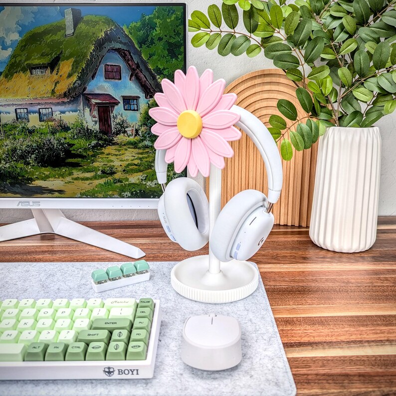 May include: White headphone stand with a pink daisy flower design. The stand is holding a pair of white headphones. The stand is on a gray desk mat with a green and white keyboard and a white mouse.