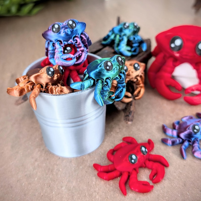 May include: A collection of colorful, hand-painted, miniature crab figurines. Some crabs are in a small silver bucket, while others are on a wooden surface. The crabs have large, expressive eyes and detailed features.