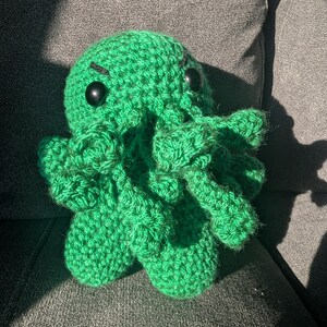 Pode incluir: Brinquedo de pelúcia verde em crochê em forma de polvo com grandes olhos pretos.