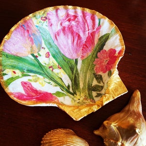 May include: A seashell dish with a floral design featuring pink tulips and green leaves. The shell is painted gold around the edges.