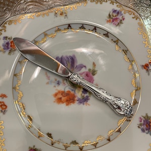 May include: A silver butter knife with an ornate handle rests on a white porcelain plate with a gold trim and floral pattern.