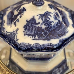 May include: A blue and white porcelain serving dish with a lid. The dish has an octagonal shape and features a detailed landscape design. The lid is topped with a small, decorative finial. The dish is sitting on a silver-colored metal tray.