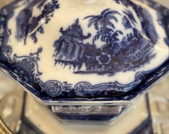 May include: A blue and white porcelain serving dish with a lid. The dish has an octagonal shape and features a detailed landscape design. The lid is topped with a small, decorative finial. The dish is sitting on a silver-coloured metal tray.
