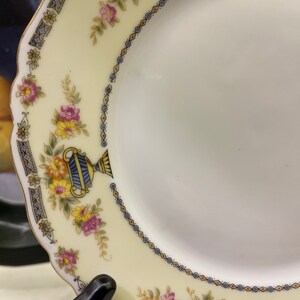 May include: A white porcelain plate with a yellow, blue, and pink floral pattern. The plate has a gold rim and a scalloped edge.