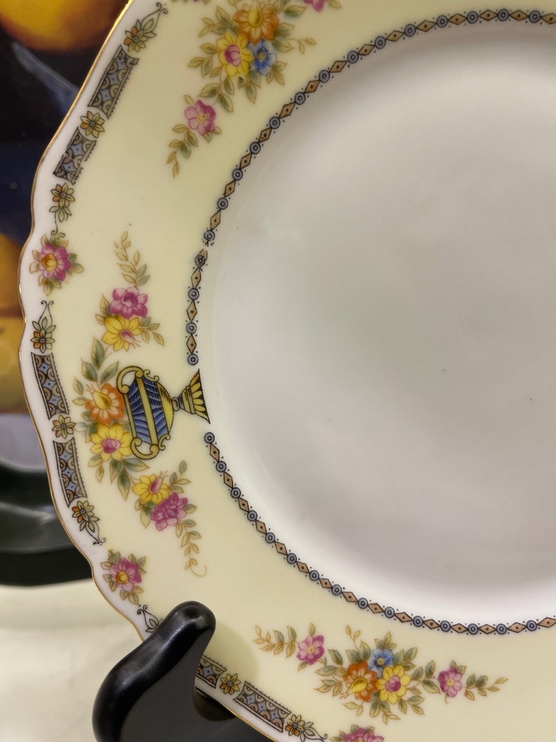 May include: A white porcelain plate with a yellow, blue, and pink floral pattern. The plate has a gold rim and a scalloped edge.