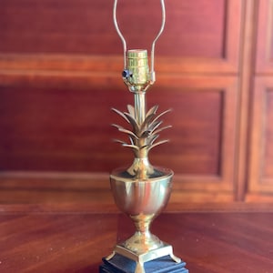 May include: A brass pineapple-shaped table lamp with a black square base. The lamp has a gold-colored stem and a bulb holder with a curved metal frame. The lamp is designed to hold a light bulb and has a power cord.