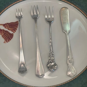 May include: Four silver utensils on a white plate with a green and gold rim. The utensils include three forks and a butter knife.