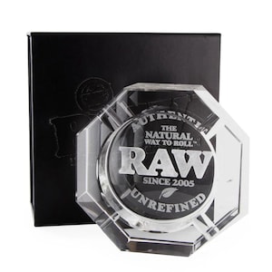 May include: A clear, octagonal ashtray with the text "RAW" and "AUTHENTIC THE NATURAL WAY TO ROLL" in silver lettering. The ashtray is set against a black box with embossed branding. The design includes the words "UNREFINED" and "SINCE 2005".