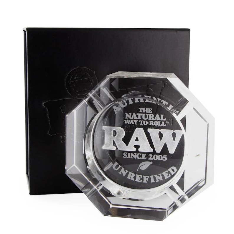 May include: A clear, octagonal ashtray with the text "RAW" and "AUTHENTIC THE NATURAL WAY TO ROLL" in silver lettering. The ashtray is set against a black box with embossed branding. The design includes the words "UNREFINED" and "SINCE 2005".
