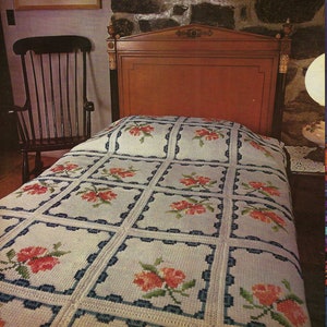 May include: A white crocheted bedspread with a floral design. The bedspread is made up of square panels with a rose and border design in each square. The bedspread is on a bed with a wooden headboard.