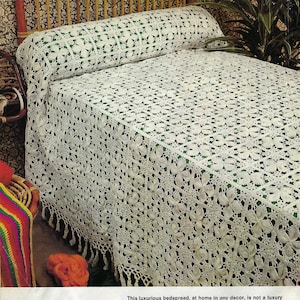 May include: A white crocheted bedspread with a floral pattern. The bedspread has a fringe border on three sides. The text in the image reads: "This luxurious bedspread, at home in any decor, is not a luxury when you make it yourself. Four ribbed leaves form the center of each square motif and a filigree of flower buds fill the corners between the leaves. Motifs are sewn together along their square border frames. Fringe bedspread on three sides for a rich finishing edge. See Trellis Bedspread, opposite page."