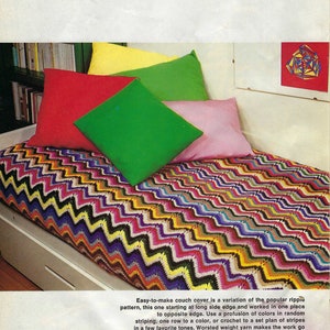 May include: A colourful crocheted couch cover with a zig-zag pattern. The cover is made with a variety of colours, including pink, purple, green, yellow, and orange. The cover is laid out on a white couch with several colourful cushions.