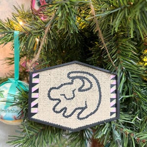 May include: A decorative Christmas ornament featuring a black and white embroidered design of a lion cub. The ornament has a black border and pink and black accents. It hangs from a natural twine string against a backdrop of a Christmas tree.