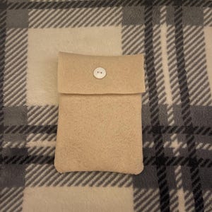 Puede incluir: Una funda de fieltro beige con solapa y cierre de bot&oacute;n blanco. La funda rectangular parece hecha a mano. El fondo es un estampado de cuadros en tonos grises y beige.