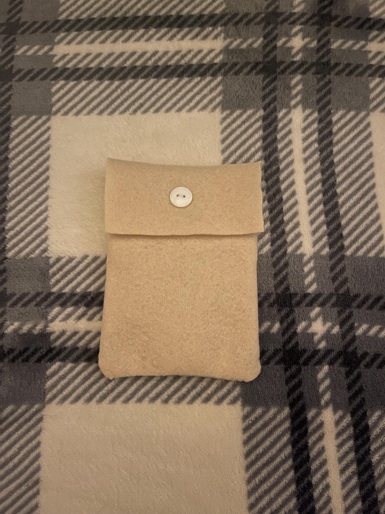 Puede incluir: Una funda de fieltro beige con solapa y cierre de bot&oacute;n blanco. La funda rectangular parece hecha a mano. El fondo es un estampado de cuadros en tonos grises y beige.