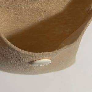 Puede incluir: Primer plano de un sombrero de fieltro beige con ala curva. Un peque&ntilde;o bot&oacute;n blanco ovalado est&aacute; cosido en el ala. El interior del sombrero es visible, mostrando la textura y el color del fieltro.