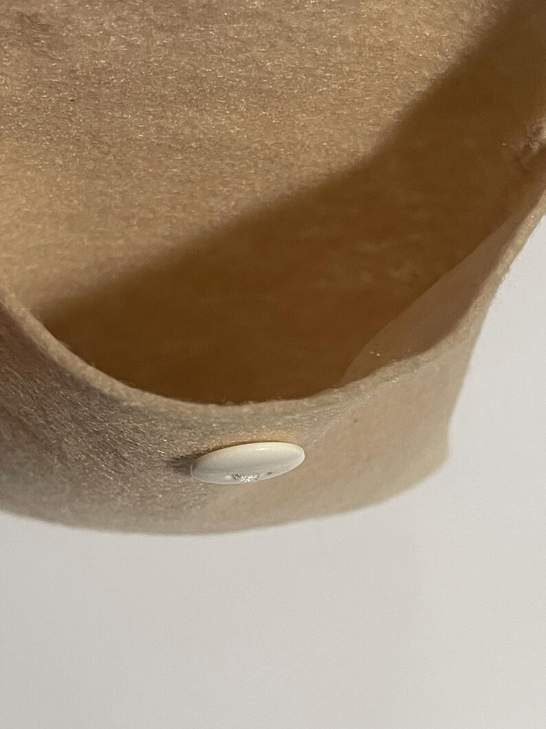 Puede incluir: Primer plano de un sombrero de fieltro beige con ala curva. Un peque&ntilde;o bot&oacute;n blanco ovalado est&aacute; cosido en el ala. El interior del sombrero es visible, mostrando la textura y el color del fieltro.