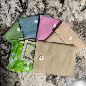 Puede incluir: Una colecci&oacute;n de bolsos de fieltro en colores pastel: verde, azul, rosa y beige. Cada bolso se asegura con un bot&oacute;n blanco. Un bolso est&aacute; abierto, revelando un billete de veinte d&oacute;lares y una compresa verde.