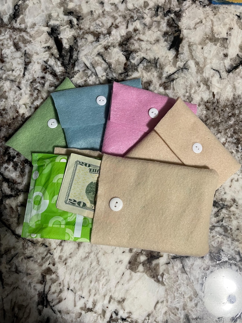 Puede incluir: Una colecci&oacute;n de bolsos de fieltro en colores pastel: verde, azul, rosa y beige. Cada bolso se asegura con un bot&oacute;n blanco. Un bolso est&aacute; abierto, revelando un billete de veinte d&oacute;lares y una compresa verde.