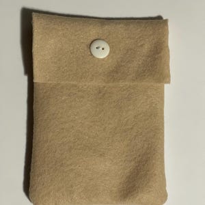 Puede incluir: Una bolsa de fieltro color beige con una solapa cerrada con un bot&oacute;n blanco. La bolsa rectangular parece hecha a mano. El fieltro tiene un aspecto suave y texturizado.